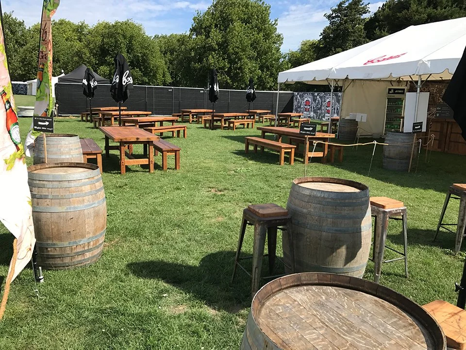 An outdoor event space with wooden picnic tables and benches, large barrels used as tables, black umbrellas, a white tent, and a grassy area with trees in the background.