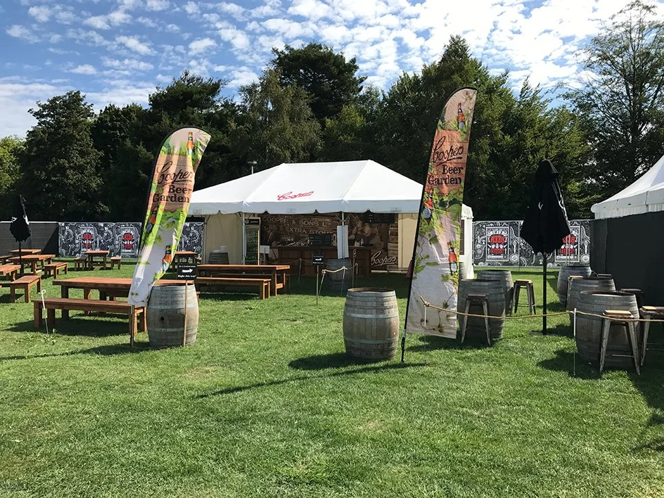 Outdoor event setup with white tent, wooden benches, barrels, flags, and tables on grass, with trees and blue sky in the background.