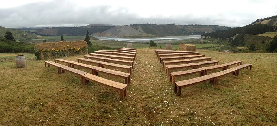 Outdoor seating with wooden benches arranged in rows on grassy terrain, overlooking a river and hilly landscape under cloudy skies.