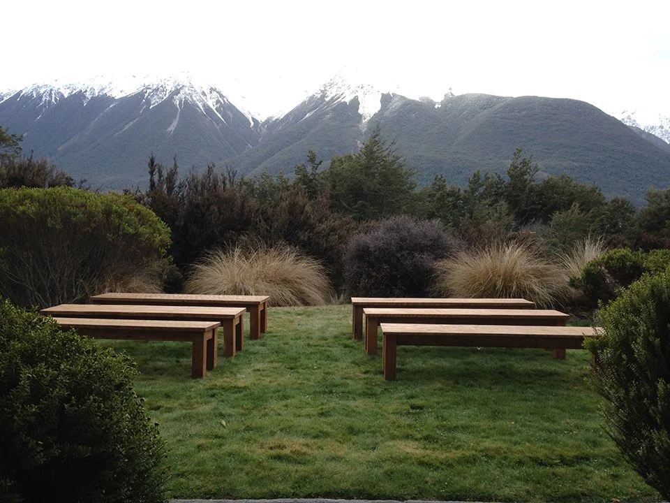 Six brown wooden benches on grass with mountain range and cloudy sky in background.