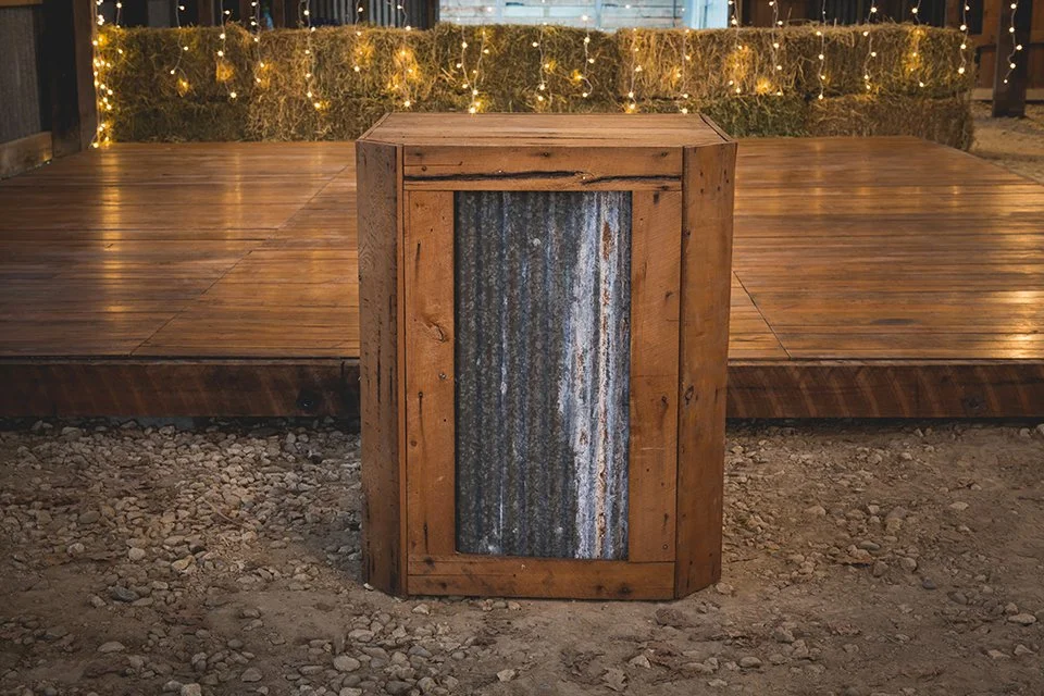 A rustic wooden trash can with a corrugated metal front, placed in front of a wooden deck with string lights hanging in the background.
