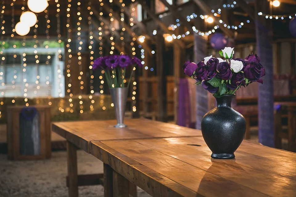 A rustic wooden table with two flower vases holding purple and white roses, set in a warmly lit indoor space with string lights and hanging paper lanterns.