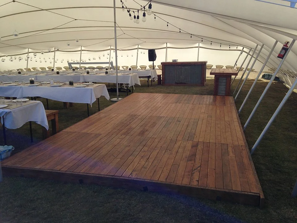 Event tent with a wooden dance floor, set up for a celebration with long tables covered in white tablecloths and chairs, string lights hanging from the tent ceiling, and a podium at the front.