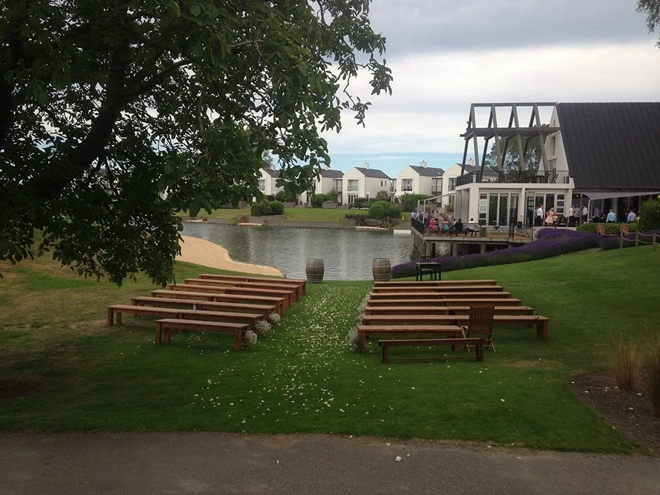 Outdoor wedding setup with wooden benches on grass, a small aisle with flower petals, a lake, and a building with a large glass window and patio where guests are gathered.