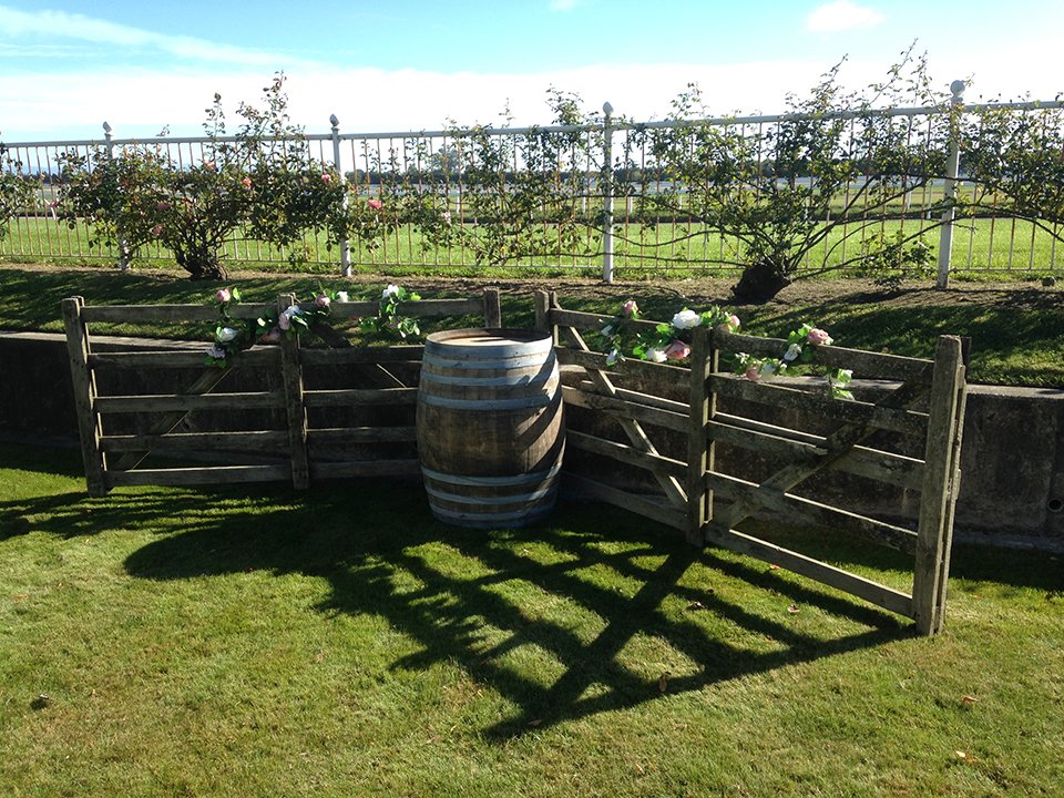 Decorative garden scene with a wooden fence, a barrel, and flower arrangements, surrounded by a grassy yard with a white fence and flowering bushes in the background.
