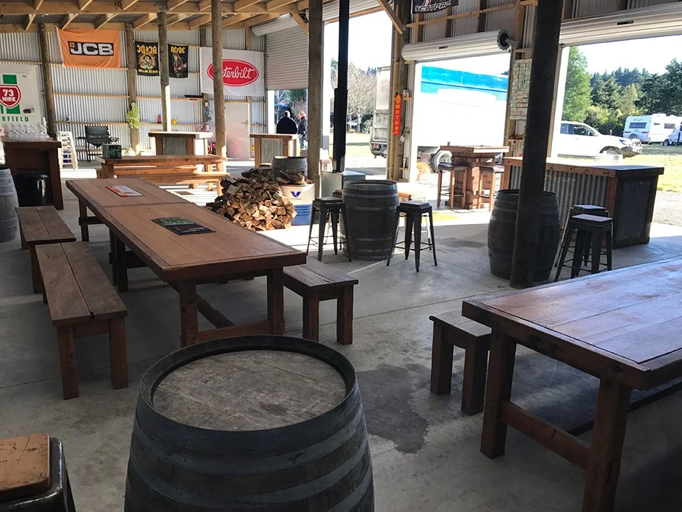 Interior of a rustic open-air restaurant or bar with wooden tables and barrel decor, sunlight streaming in from open doors, and firewood stacked in the center.