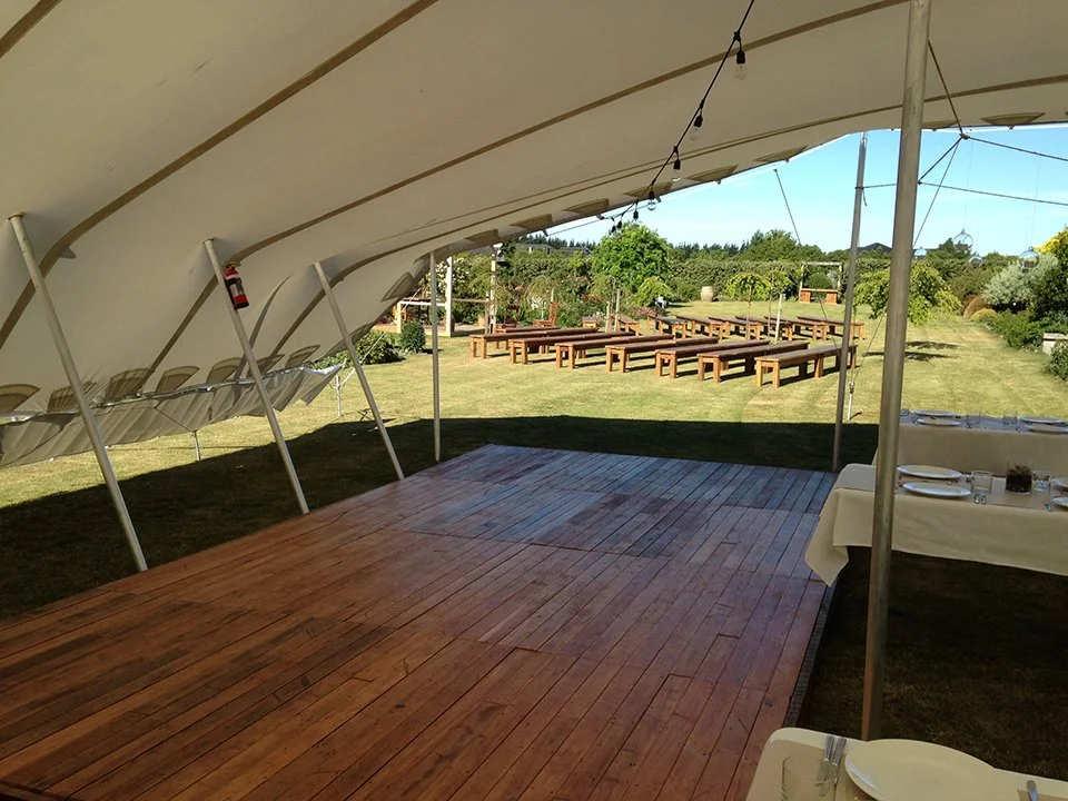 An outdoor event space with a large white tent, a wooden dance floor, and seating tables with plates and cutlery, set in a garden with trees and greenery in the background.