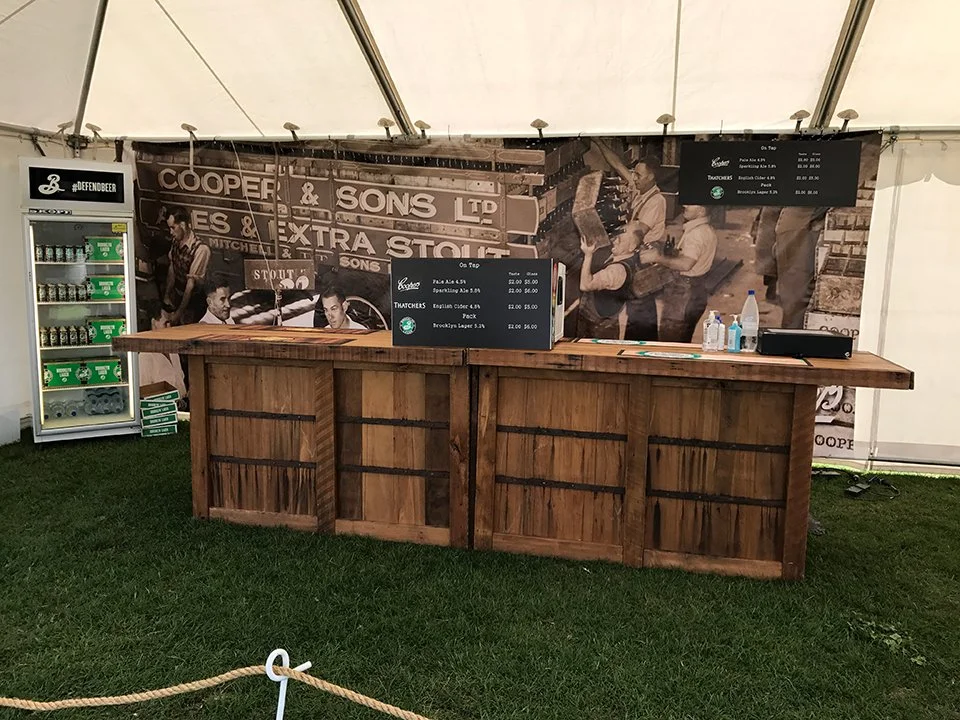 A wooden bar counter inside a tent with drink menus displayed on a small blackboard and a larger digital menu. Bottles, hand sanitizer, and a water bottle are on the counter. A vintage black-and-white wallpaper with historical images and text is in t