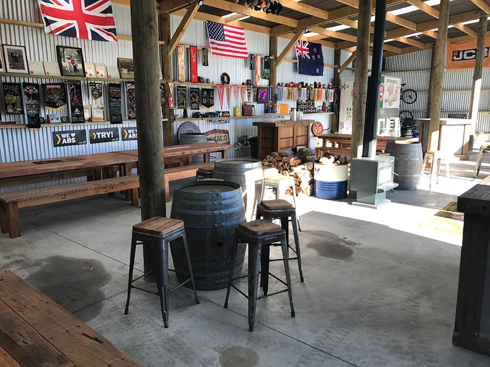 Interior of a rustic bar or gathering space decorated with U.S. and Australian flags, featuring wooden tables, barrels, and assorted memorabilia on the walls.
