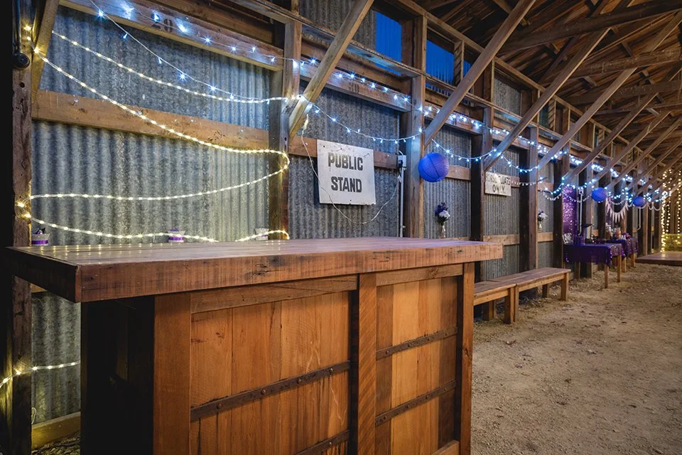 Decorated rustic barn interior with string lights, paper lanterns, and a sign that says 'Public Stand', featuring a long wooden counter and benches for guests.