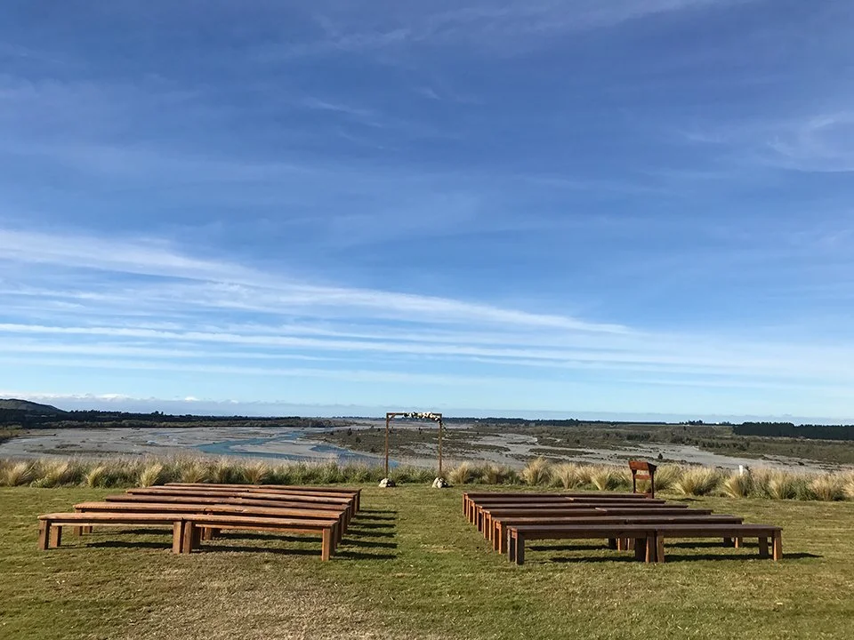 Empty wooden benches on a grassy area facing a river and landscape with a bright blue sky and sparse clouds.