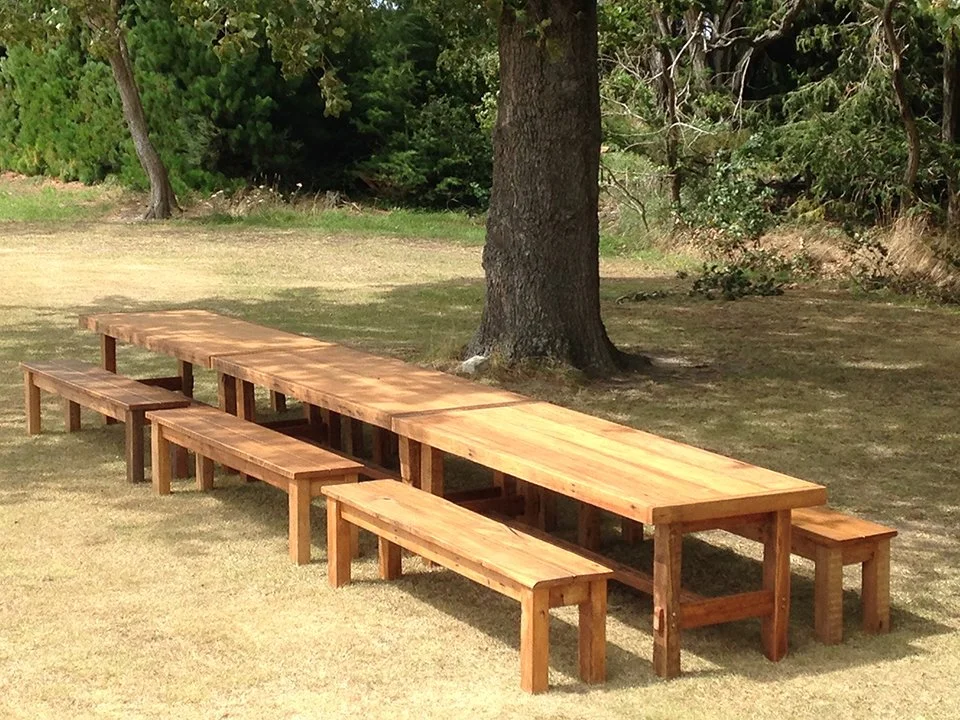 A set of wooden picnic tables with attached benches on a grassy area with trees in the background.