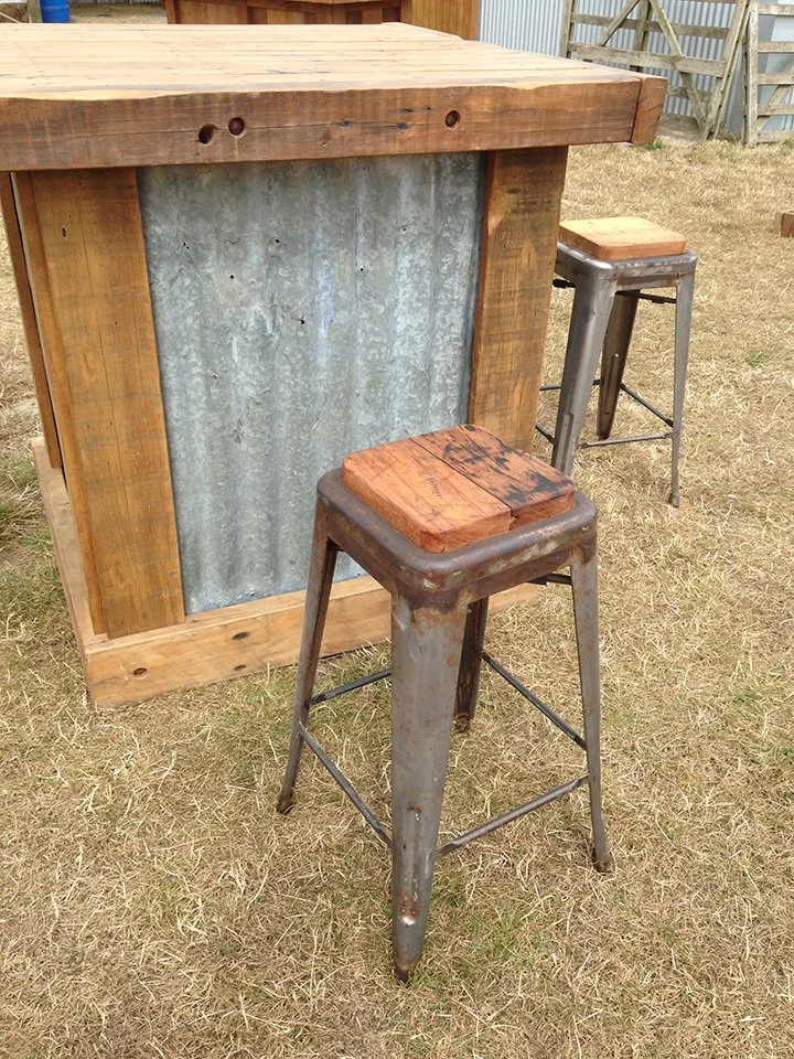 An outdoor scene featuring a rustic wooden table with a corrugated metal panel on the front, and two metal bar stools with wooden tops on dry grass.