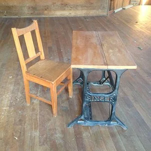 A vintage wooden sewing machine table repurposed as a desk with a wooden chair placed next to it on a hardwood floor.