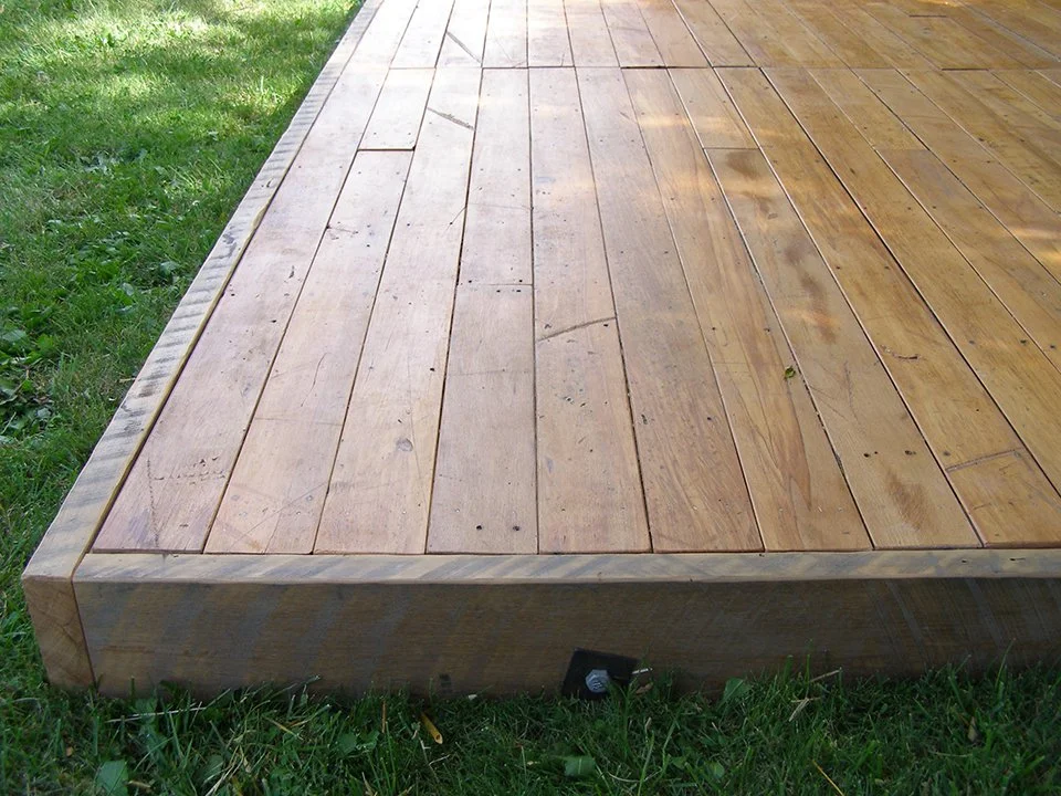 A wooden deck with a border around the edges, built on grass with some greenery nearby.