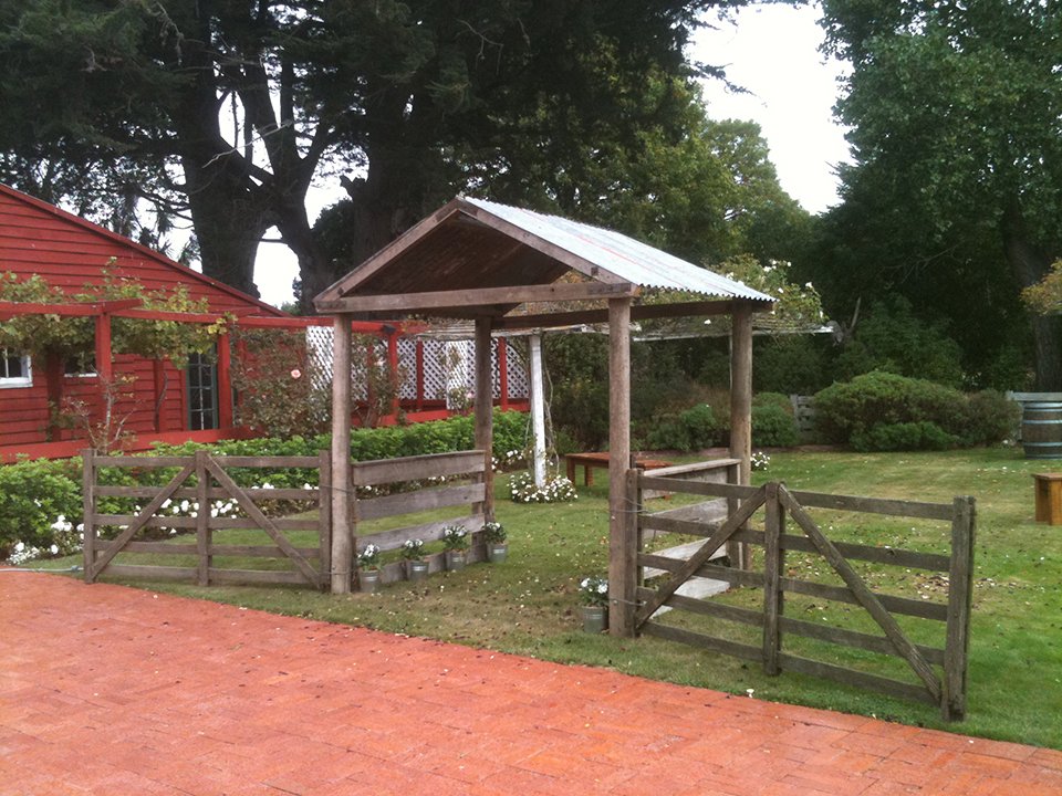 A small wooden garden gazebo with a metal roof, surrounded by a wooden fence and located on a grassy area with trees and shrubs. There is a red building behind with some windows and white latticework. The scene appears to be part of a park or backyar