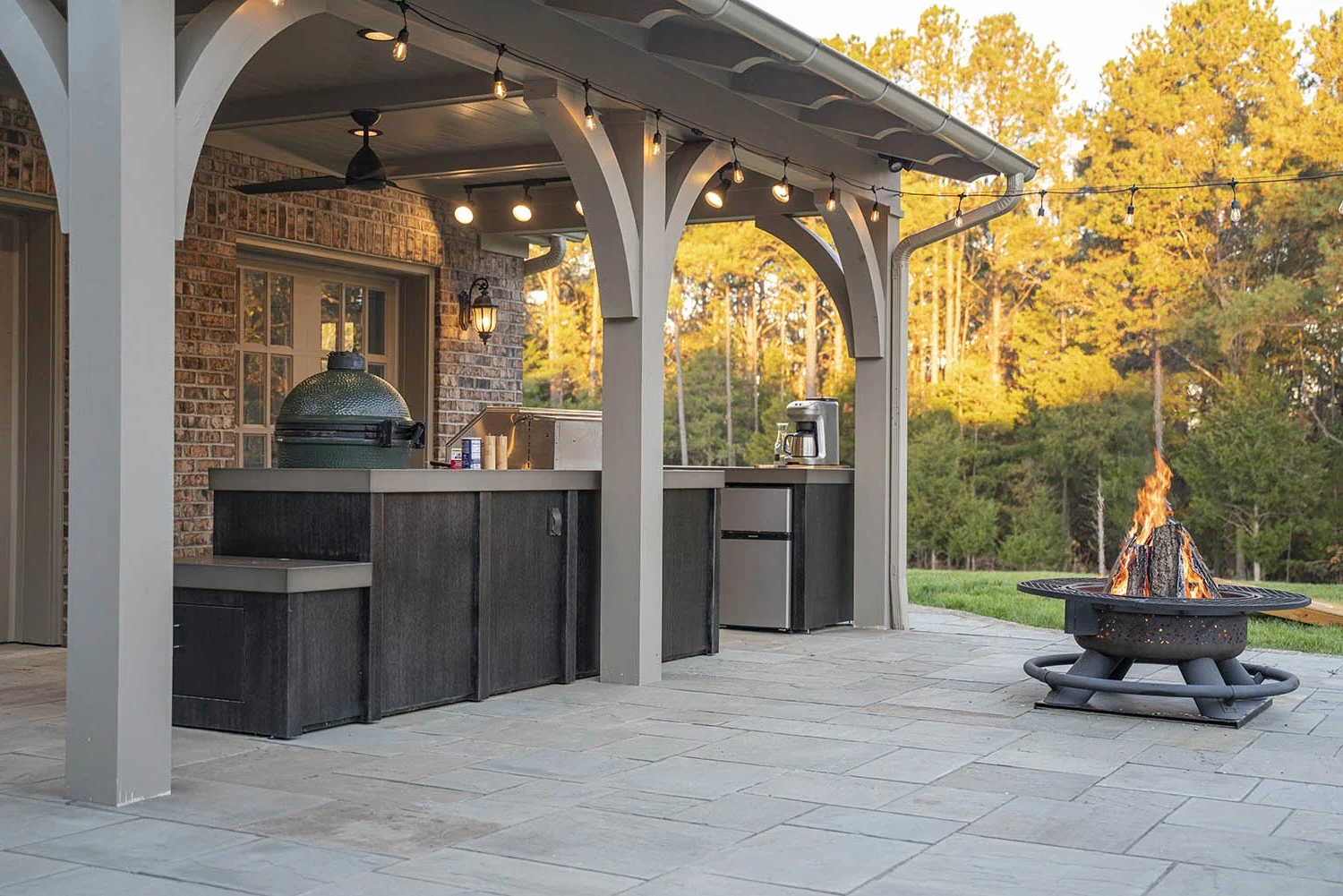 Exterior patio with a firepit, kitchen island, and string lights, surrounded by trees with fall foliage in the background.