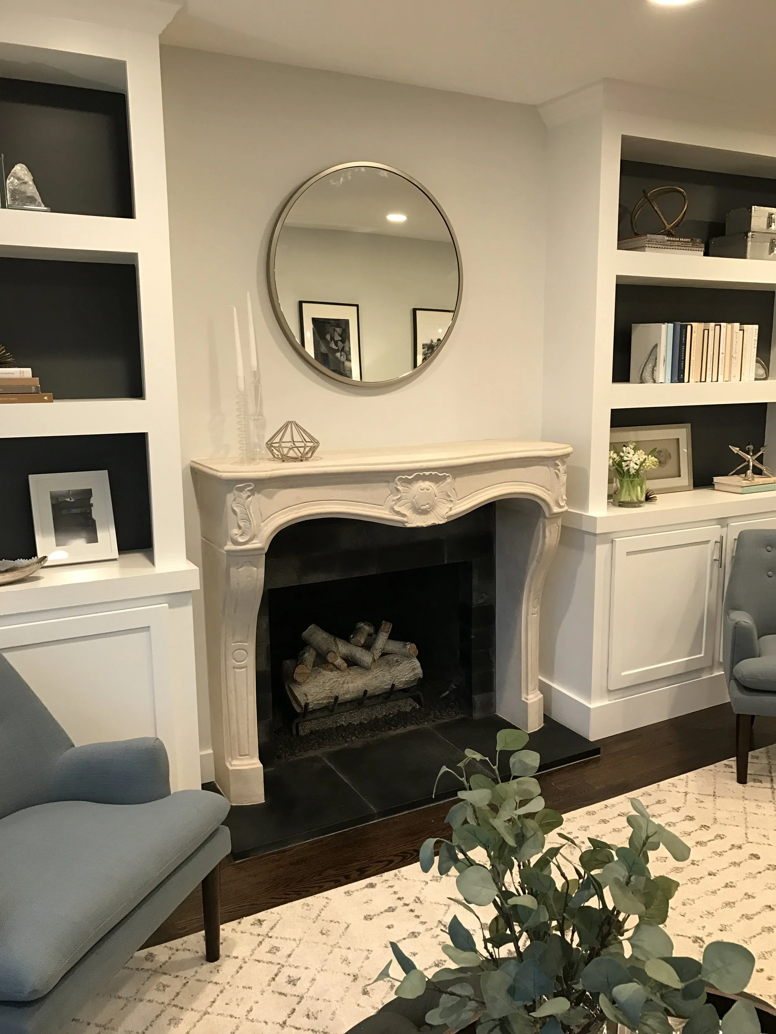 Living room with a white ornate fireplace, a round mirror above it, built-in bookshelves on both sides, a light blue armchair, and a patterned rug.