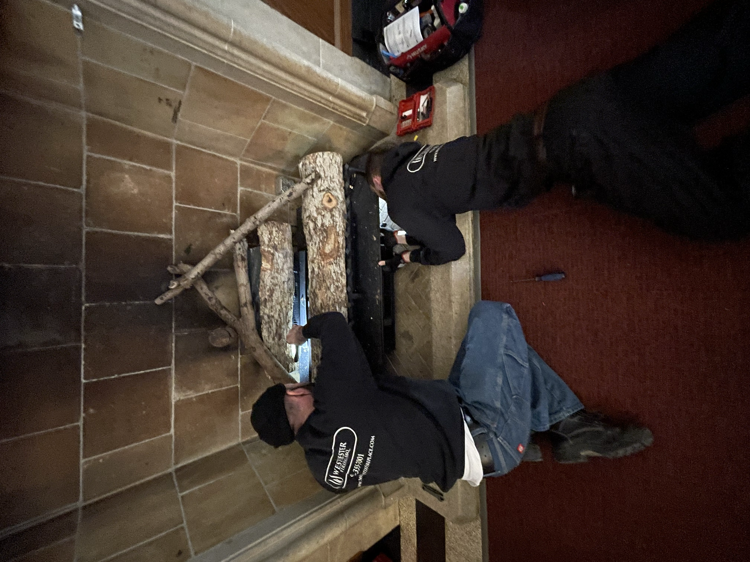 Two men installing a fireplace surround with logs inside. One man is kneeling, working at the bottom of the fireplace, while the other man is standing and assisting.