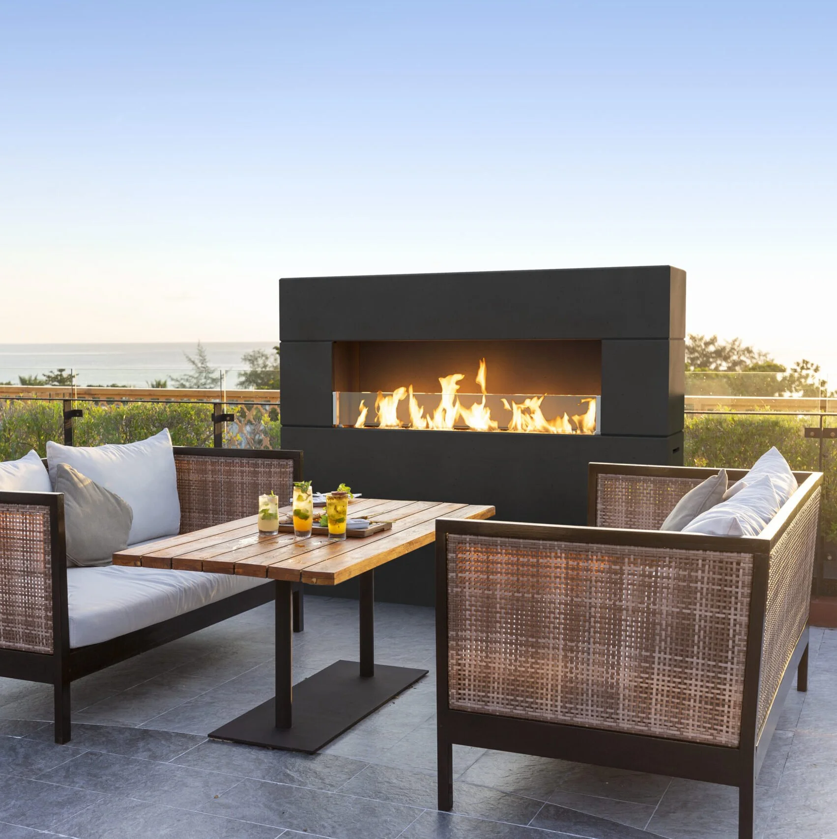 Terrace outdoor seating area with a wooden table, white cushioned chairs, drinks, and a modern rectangular fireplace with flames, overlooking a landscape with trees and a body of water in the distance.