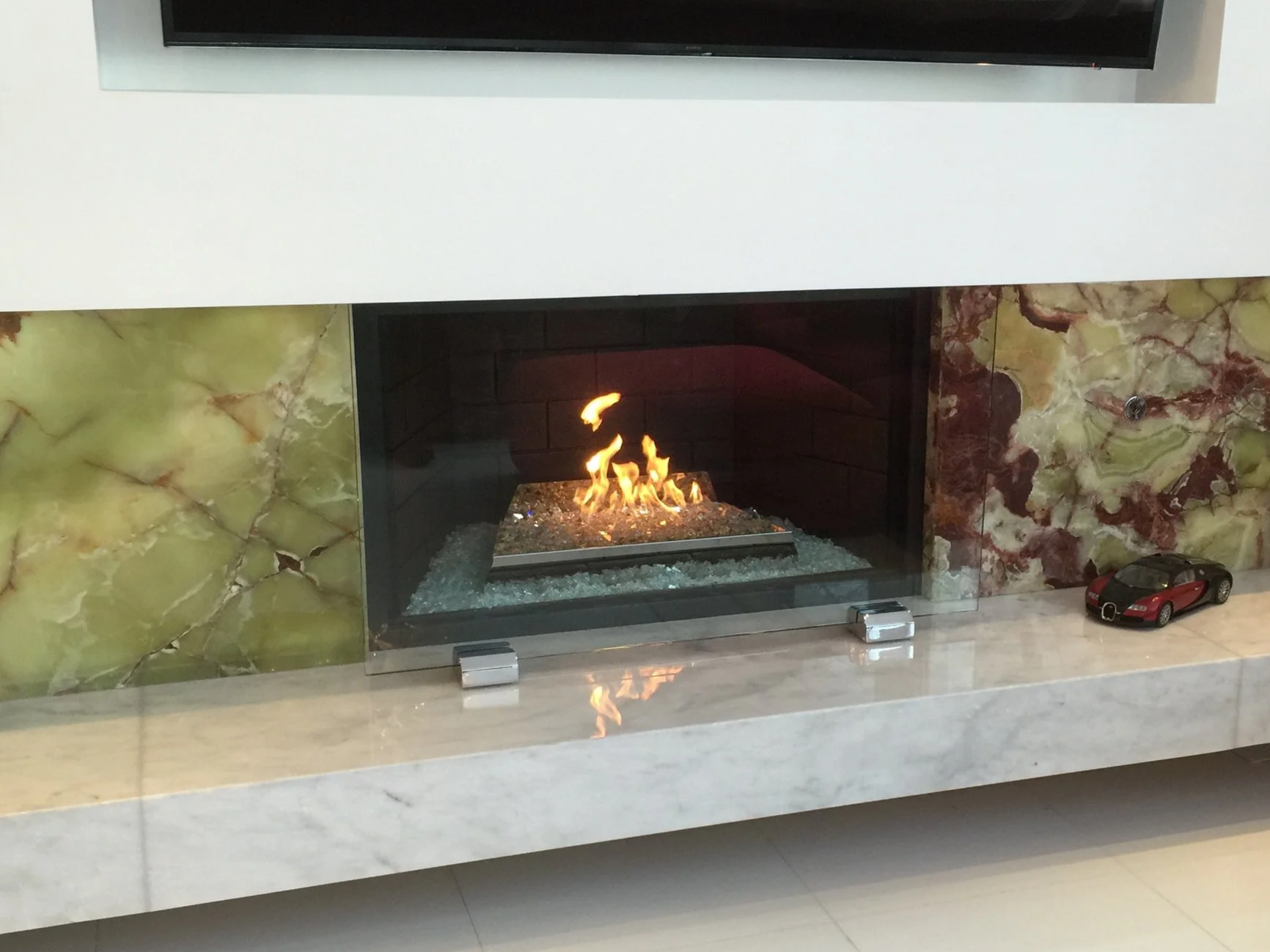 Modern fireplace with flames burning on a bed of glass stones, surrounded by a marble mantel with a small toy car on the right.