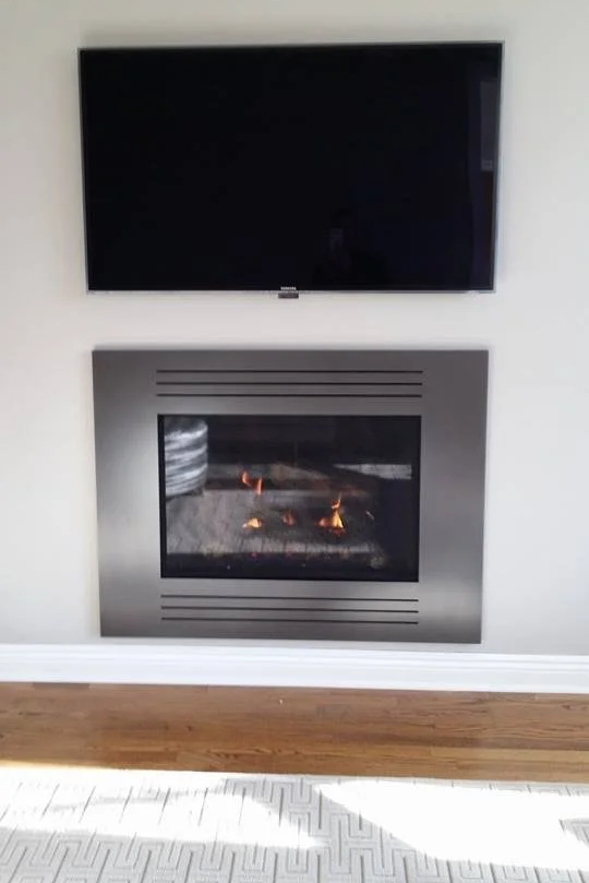 A flat-screen TV mounted on the wall above a modern fireplace with a glass front showing flickering flames, in a room with light-colored walls and wooden flooring.