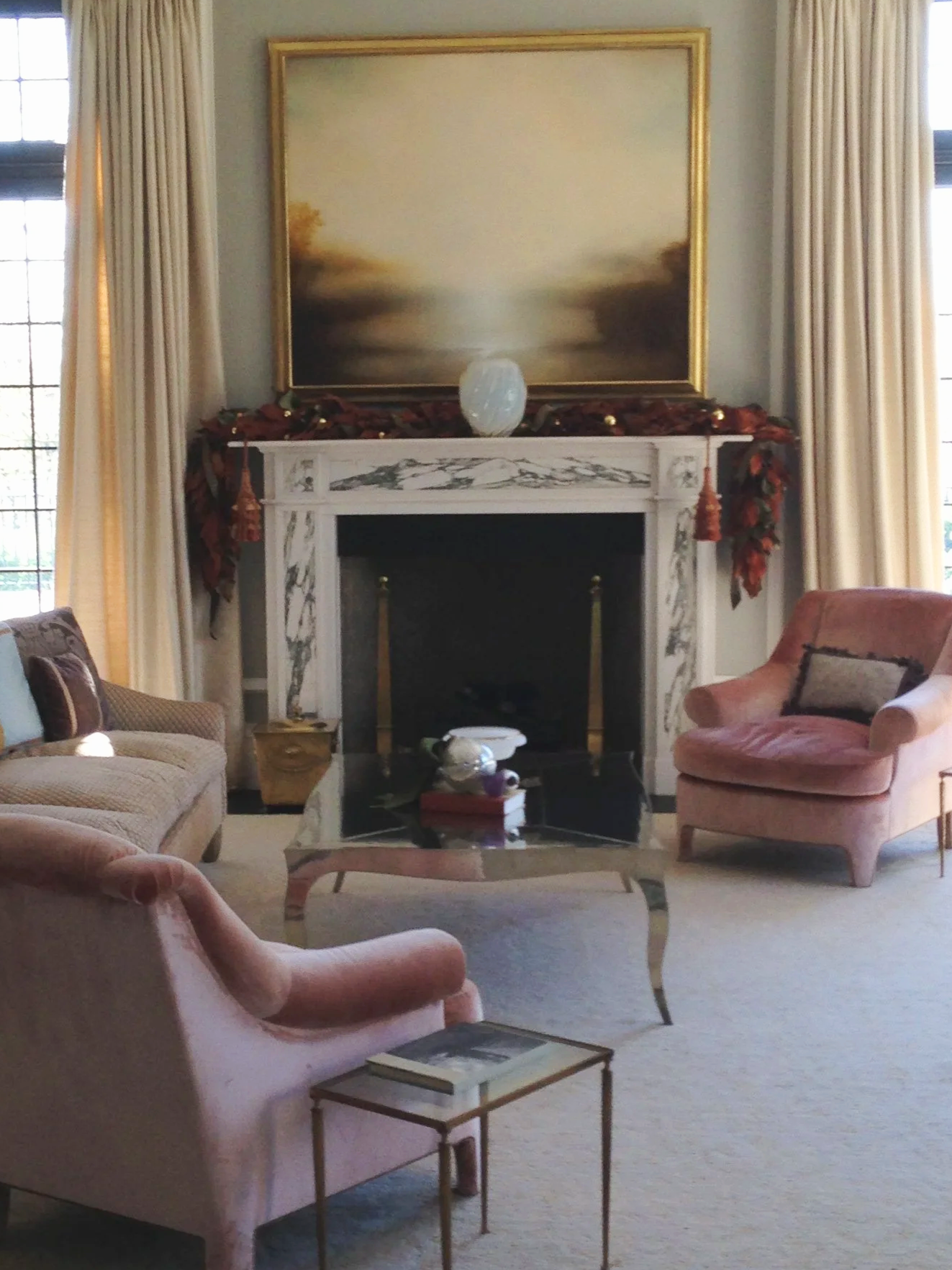 Elegant living room with a pink armchair, a beige sofa, a glass coffee table, a marble fireplace, a large abstract painting, and cream curtains.