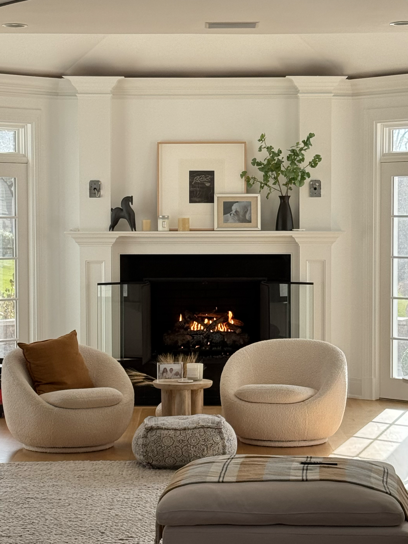 Living room with a fireplace, two cream-colored armchairs, a round wooden side table with framed photos and candles, a patterned ottoman, and a beige area rug. Decor on the mantle includes framed artwork, a black horse figurine, candles, and a vase w