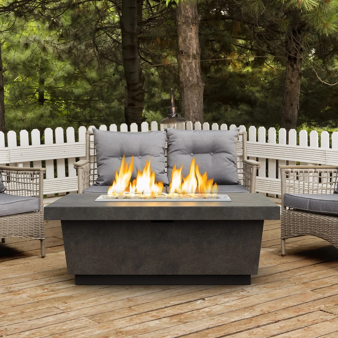 Outdoor patio with a fire feature, cushioned wicker chairs, white picket fence, trees, and a wooden deck.