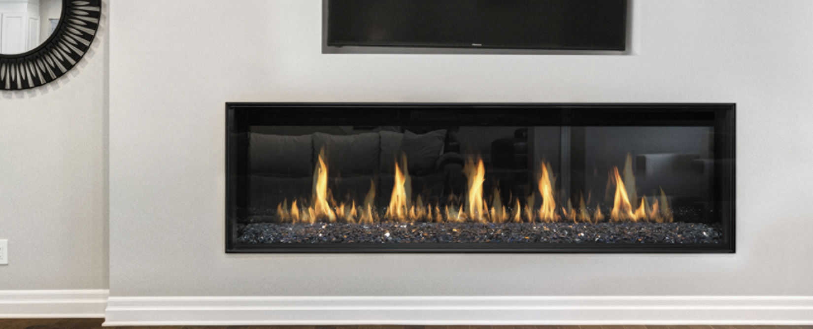 Modern linear fireplace with flames and decorative black glass stones, installed in a light-colored wall, with a TV mounted above and a mirror on the adjacent wall.