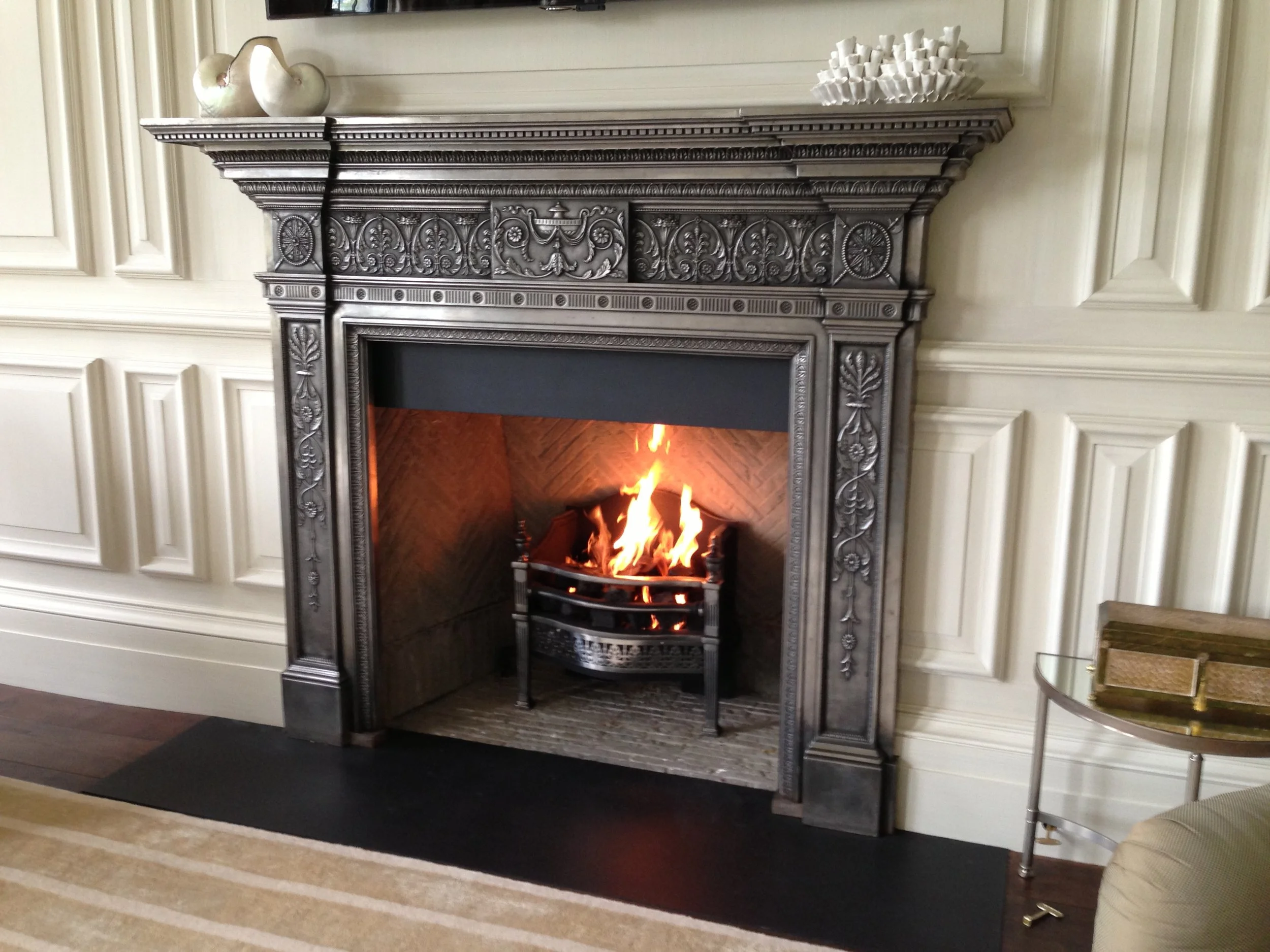 A decorative fireplace with a burning fire inside, framed by ornate dark wood with carved floral patterns, in a room with cream-colored paneled walls, a black hearth, and a small side table with a glass top.