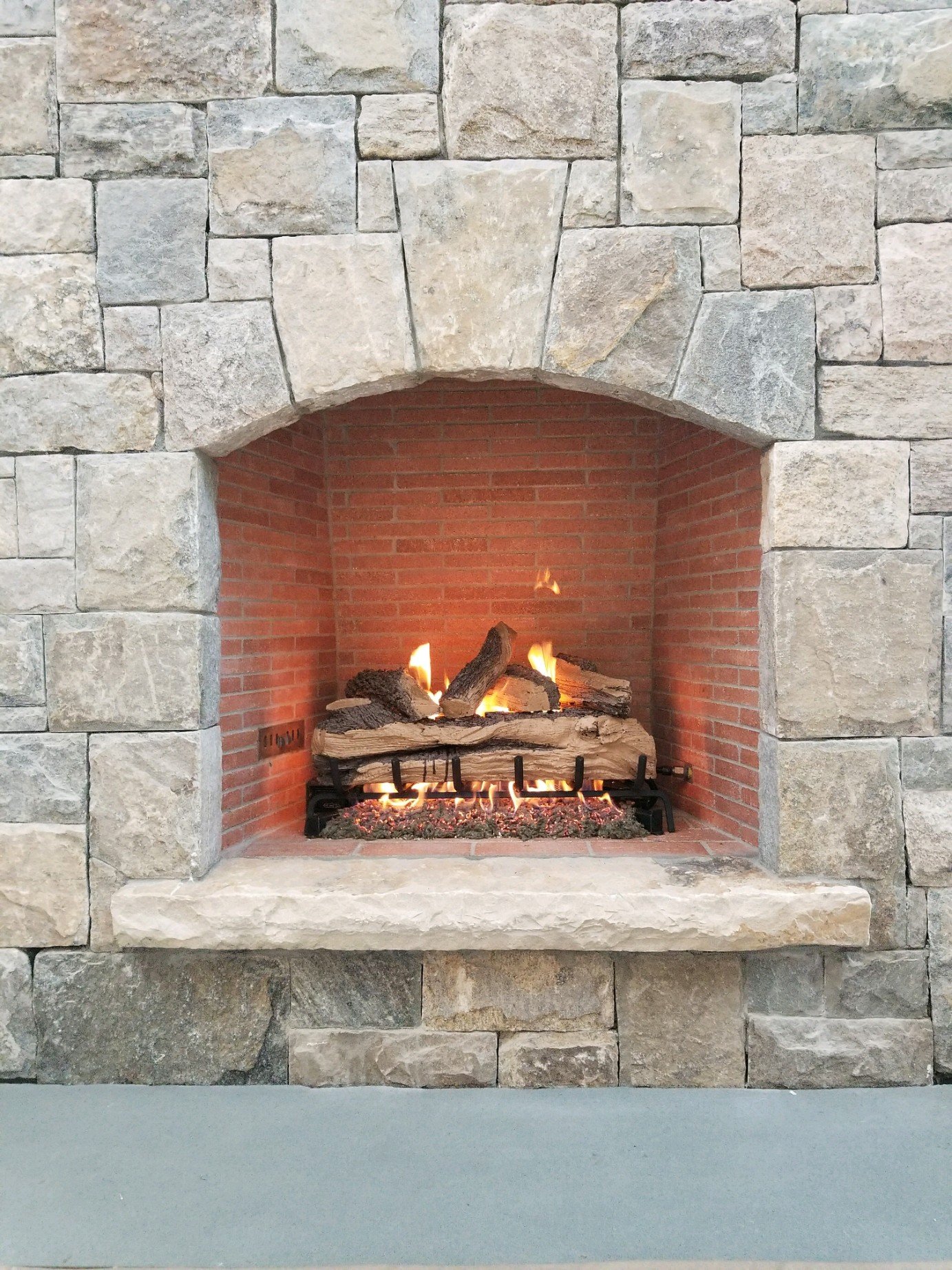 A cozy fireplace with a stone exterior and a brick interior, featuring a gas fire with logs and flames.