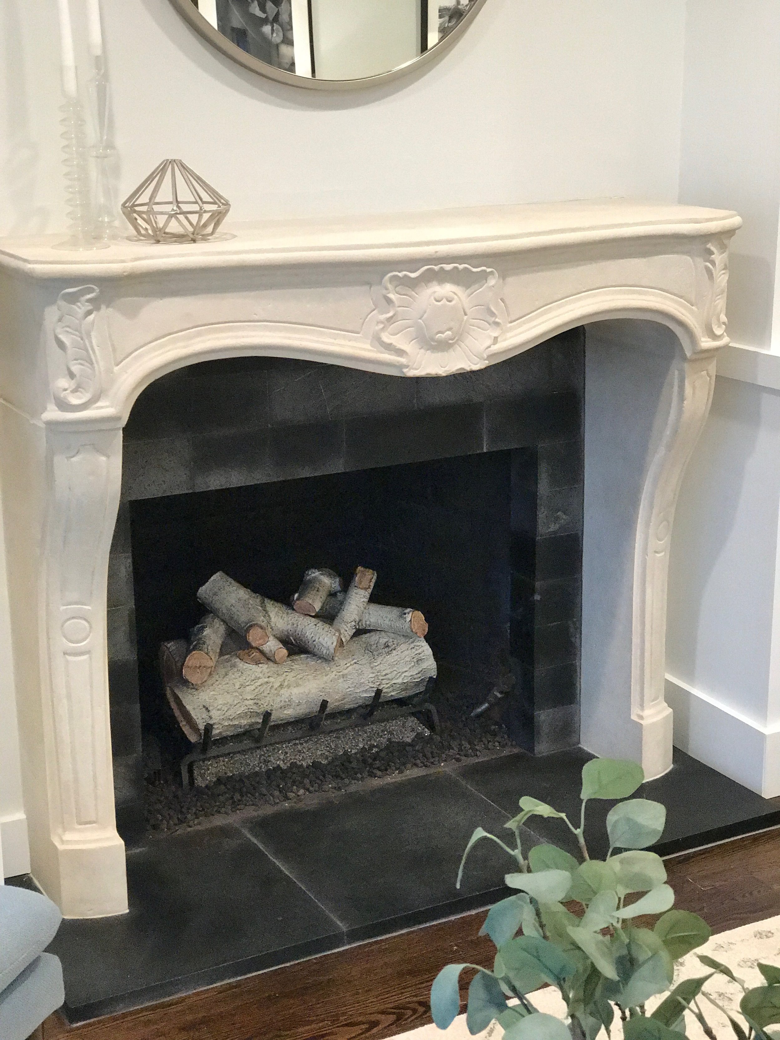A decorative white fireplace mantel with carved lion face detail, containing stacked firewood logs inside, in a living room with a mirror above and some decorative items on top.