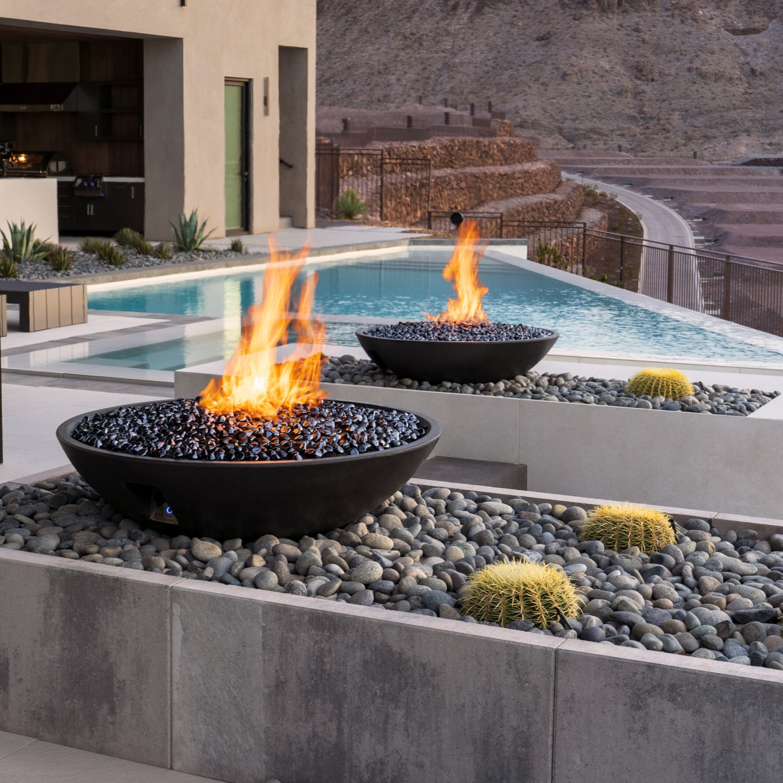 Modern backyard with swimming pool, fire pits with flames, and desert landscape with cacti and rocky terrain.