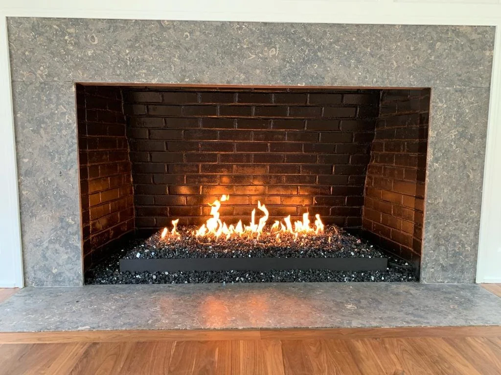Modern fireplace with a fire burning on black volcanic rocks, surrounded by a stone and brick mantle, in a living room with wooden flooring.