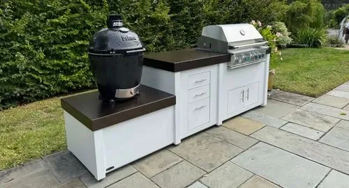 Outdoor kitchen with a black smoker on the left, a black countertop, and a white grill in the center on a concrete patio with a grassy yard and trees in the background.