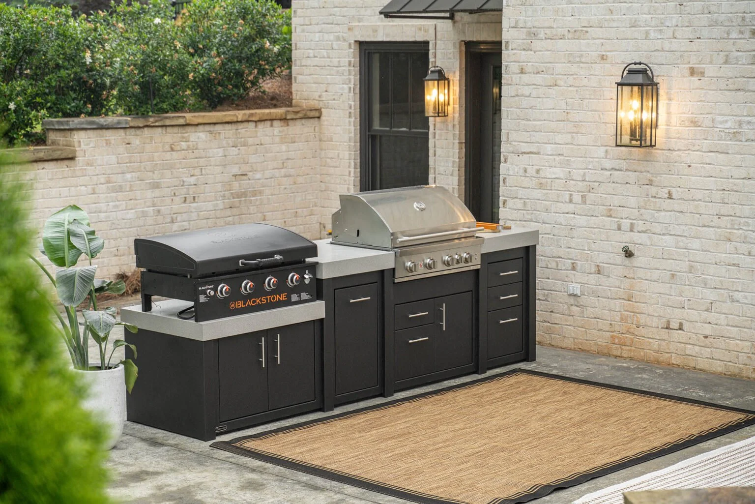 Outdoor kitchen area on a patio with a black gas grill and a stainless steel grill laid out on black cabinets with silver handles, against a brick wall with two outdoor wall lamps, a sliding glass door, and a beige outdoor rug on concrete flooring.