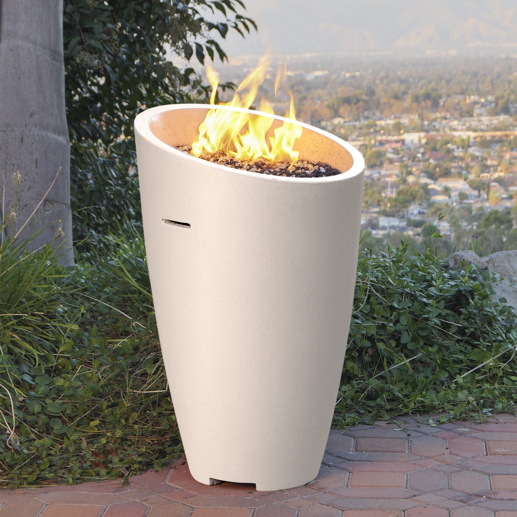A modern outdoor fire pit in a tall, white, oval-shaped design with a fire burning inside. The fire pit is placed on a brick patio surrounded by greenery, overlooking a cityscape in the background.