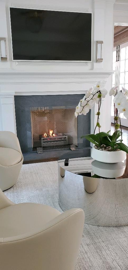 Living room with a flat-screen TV above a fireplace, a white orchid plant on a glass coffee table, and cream-colored armchairs.