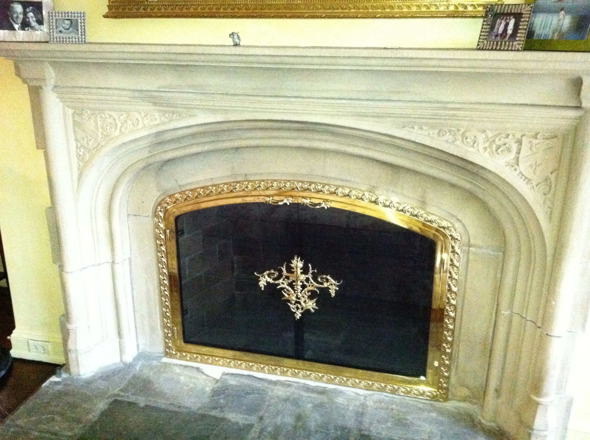 A decorative fireplace with an ornate white marble mantel, gold trim, and black interior with a gold decorative element hanging in the middle.