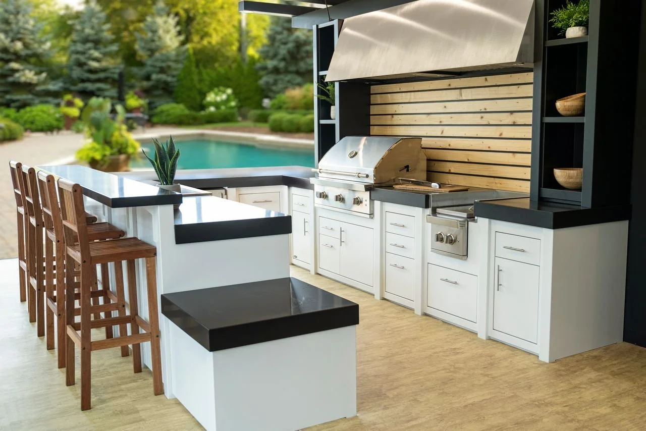 Outdoor kitchen with white cabinets and black countertops, built-in grill, and a view of a swimming pool and lush greenery in the background.