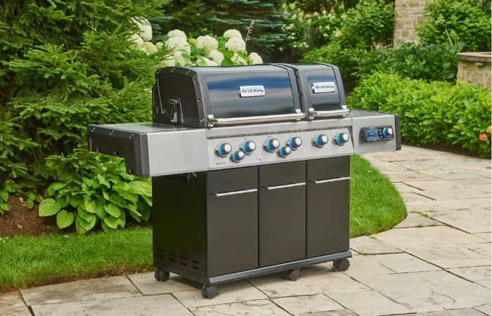 A black and gray outdoor gas barbecue grill with side tables and control knobs, situated on a stone patio in a garden setting.