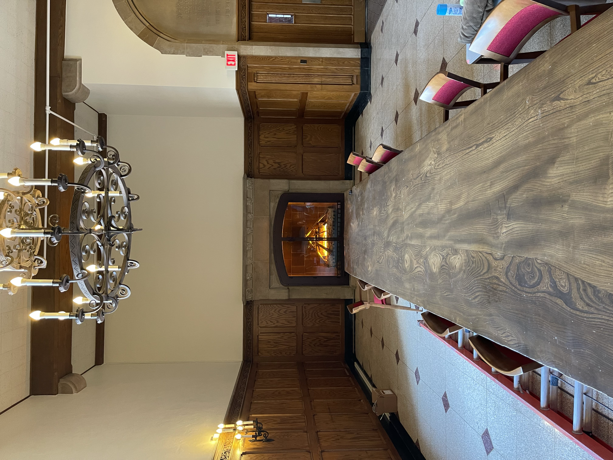 An elegant dining room with a long wooden table, red cushioned chairs, a stone fireplace with a fire burning, wooden paneling on the walls, and a chandelier hanging from the ceiling.