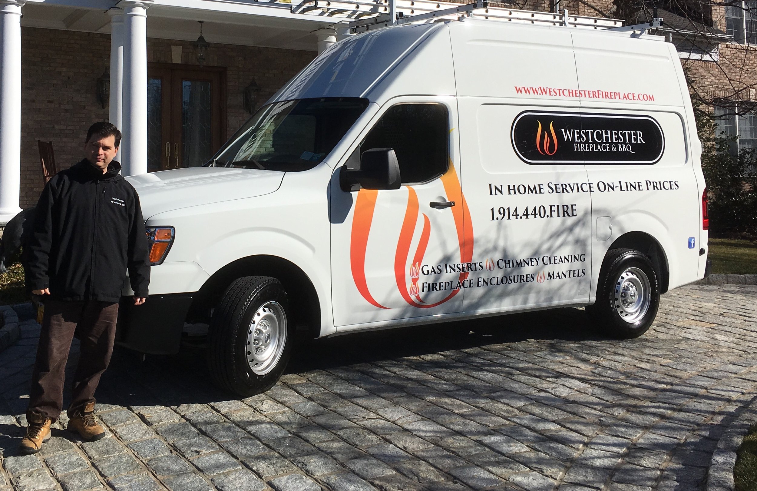 A man standing next to a white van with branding for Westchester Fireplace & BBQ parked on a cobblestone driveway in front of a house.