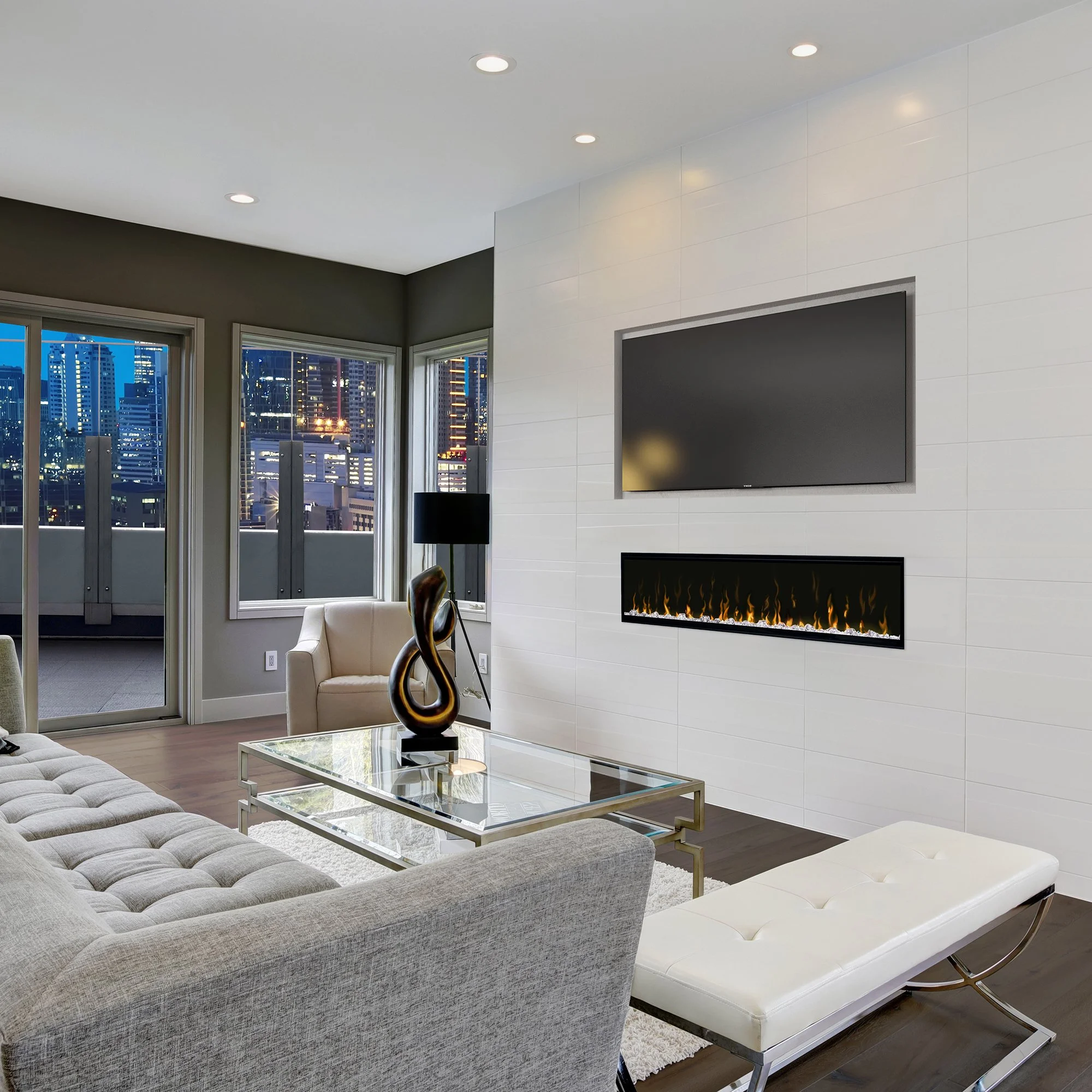 Modern living room with a beige couch, glass-top coffee table, and an abstract sculpture. Wall-mounted TV and linear fireplace on white tiled wall. Large windows showing a city skyline at dusk.