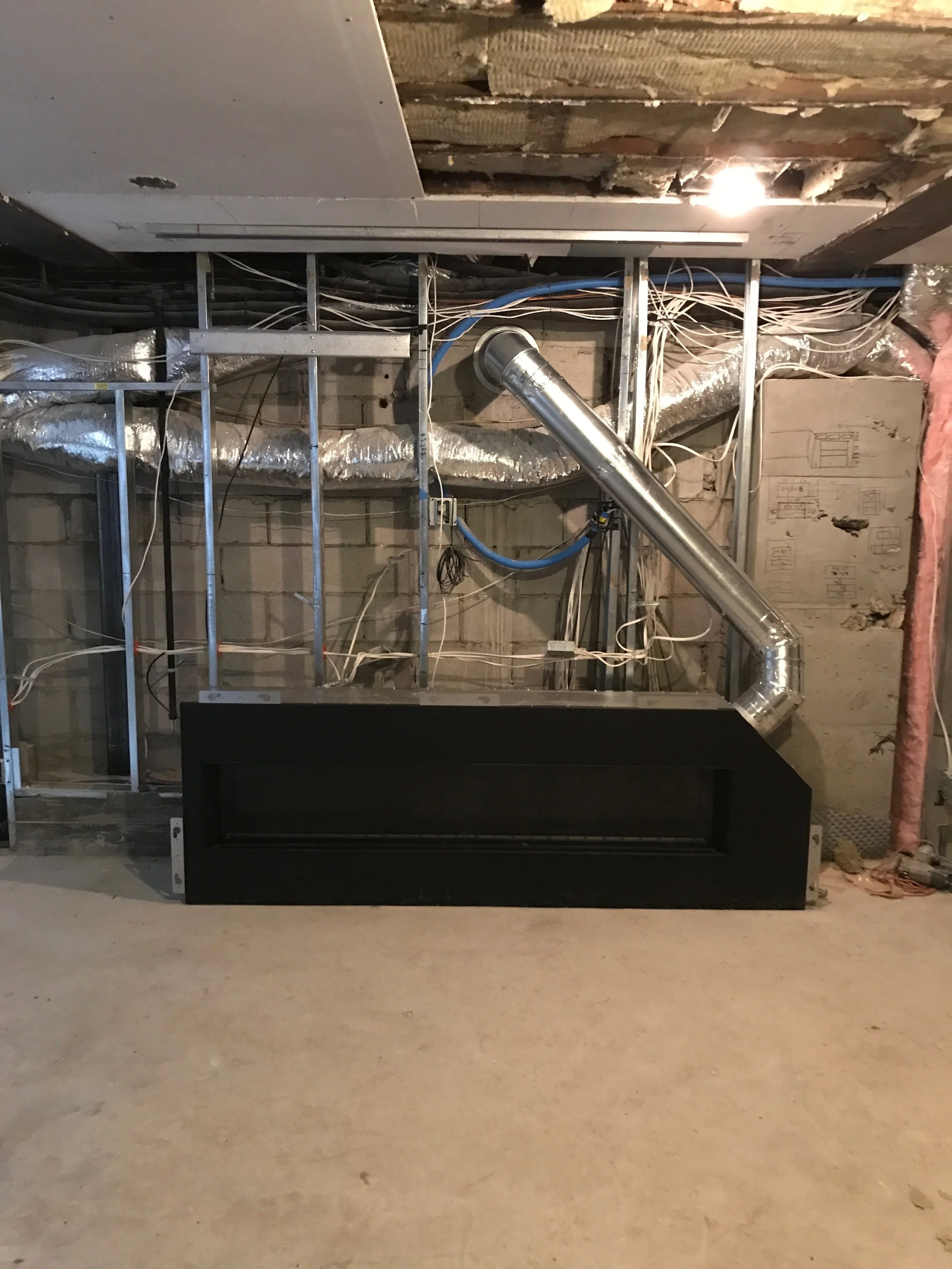Construction site showing exposed metal framing, electrical wires, and ductwork with a black box or vent in the foreground, and a partially finished ceiling with insulation on the right.