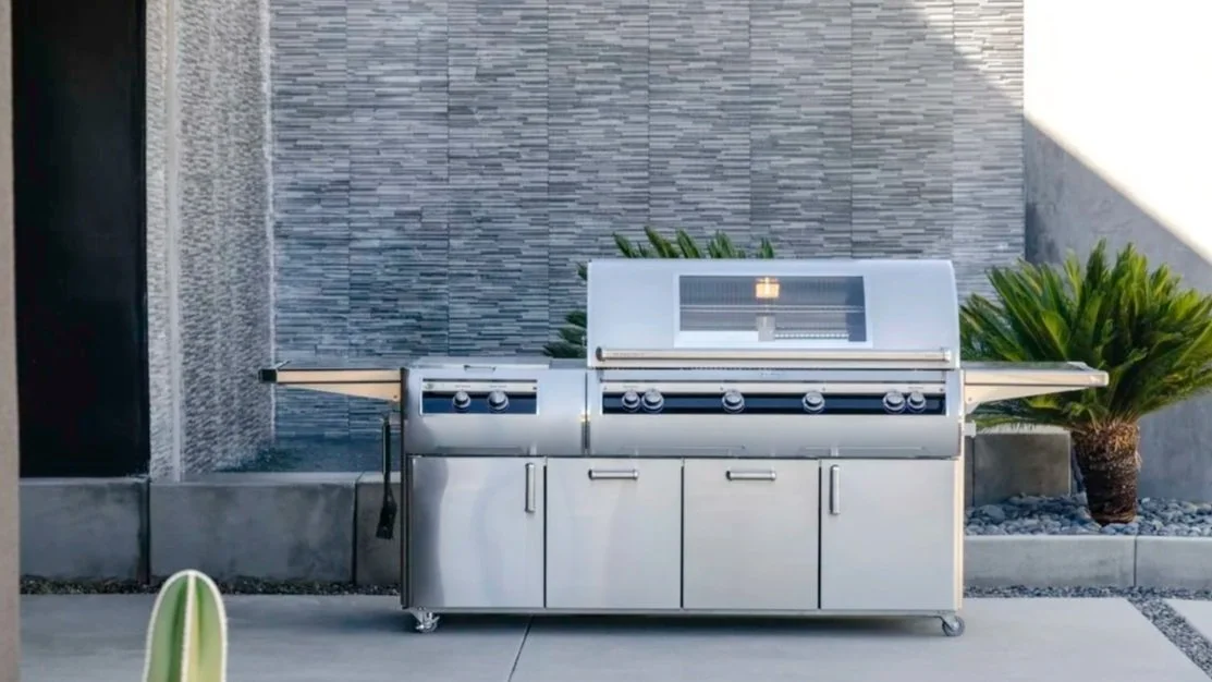 A stainless steel outdoor grill on a concrete patio with a decorative stone wall, greenery, and a small plant in the foreground.