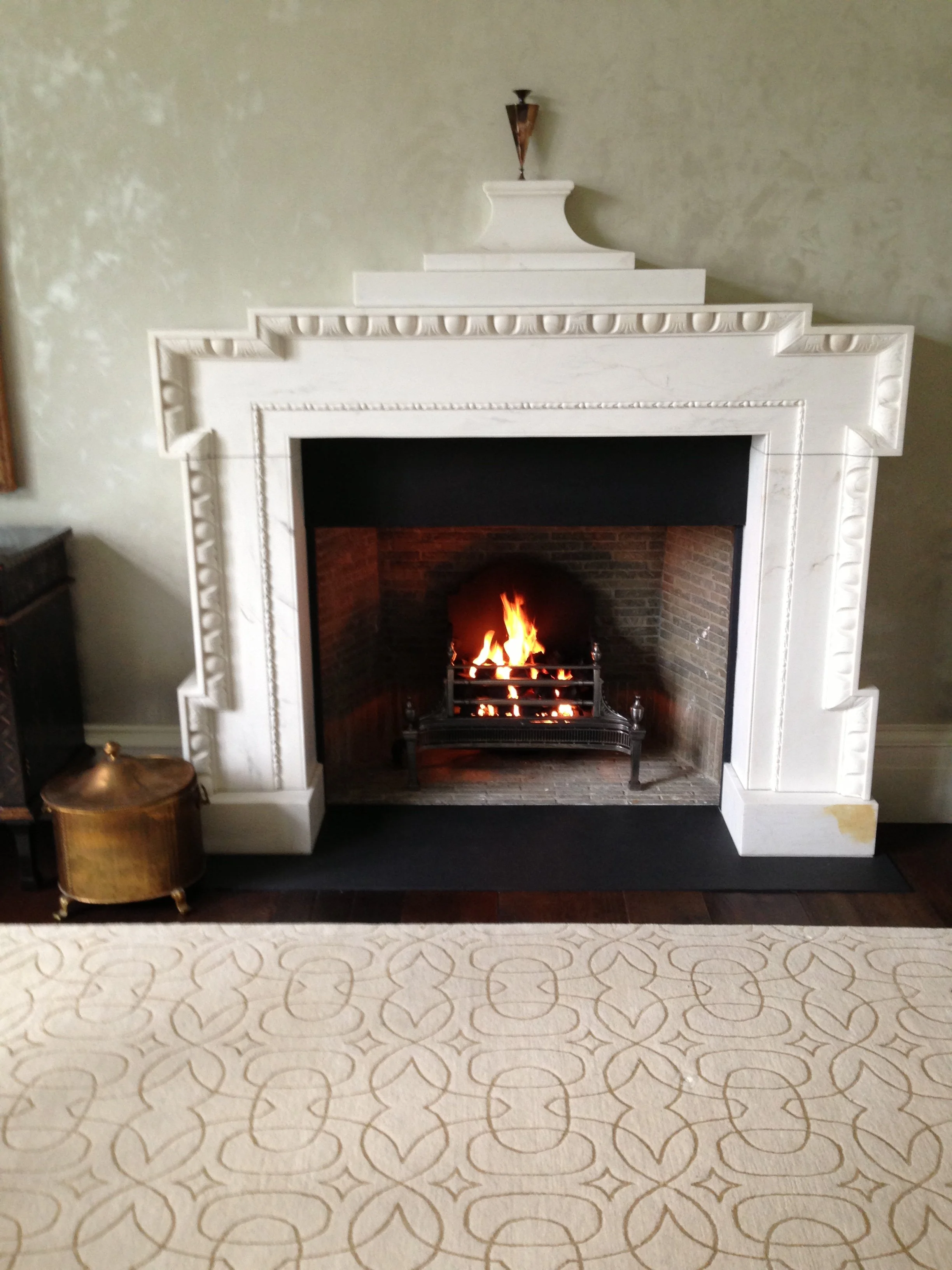 White fireplace with ornate molding, a fire burning inside, and decorative objects on top, set in a living room with patterned carpet and beige walls.