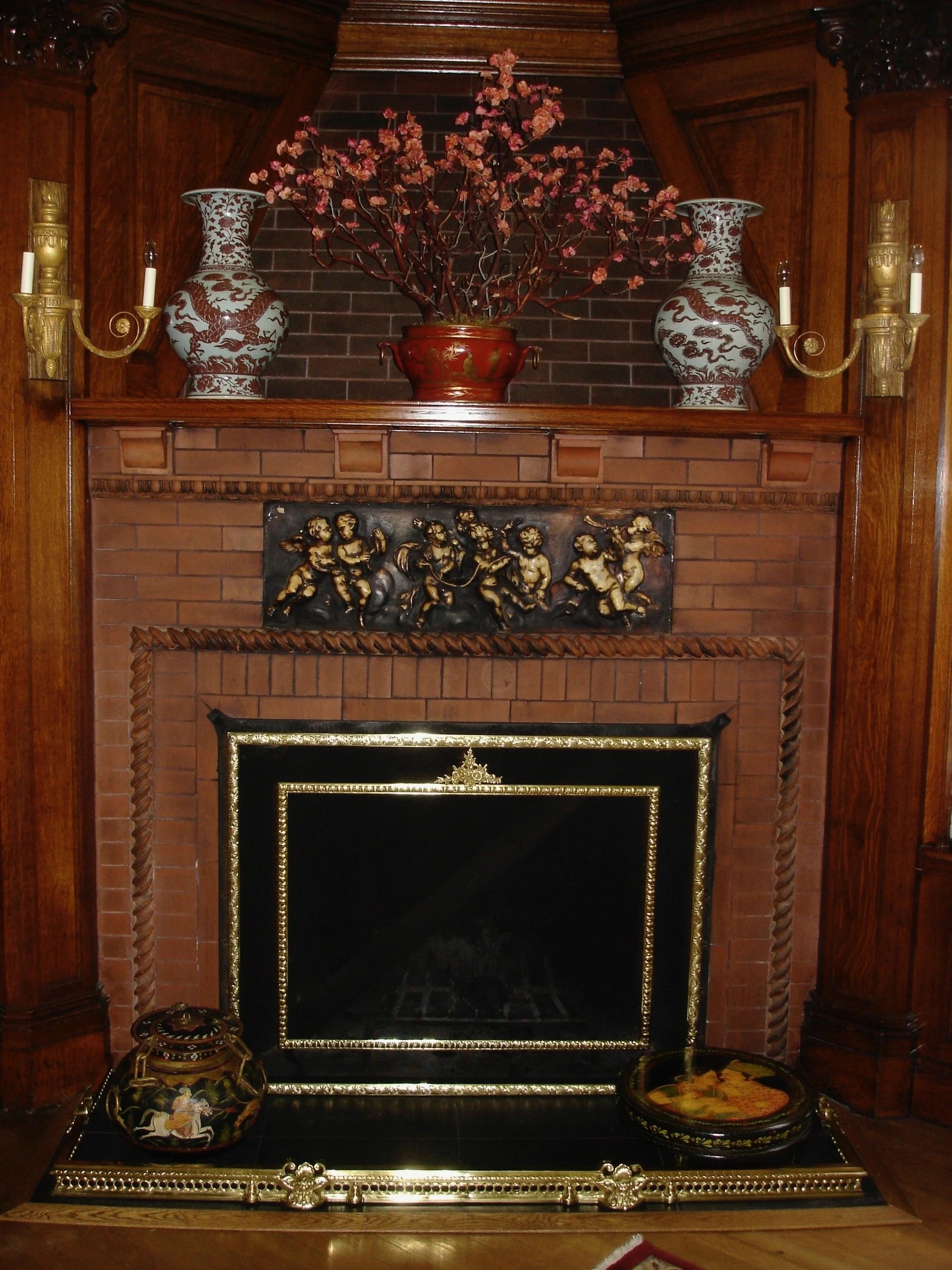 A decorated fireplace with ornate vases, a branch with pink blossoms, a carved bronze panel, and decorative items on the hearth.