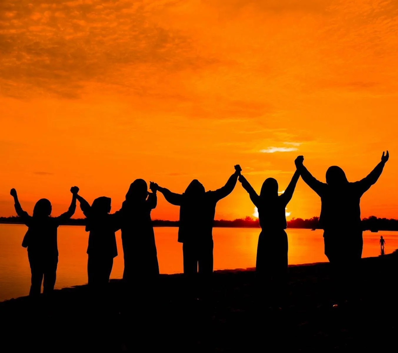 silhouetted group of people holding hands with arms raised looking at brilliant orange sunset